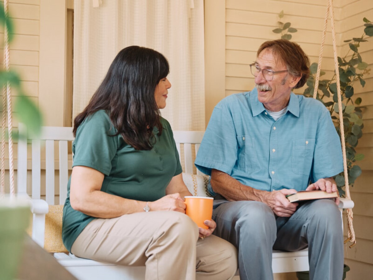 A Home Instead caregiver sits on a porch swing with an elderly man, both smiling warmly, highlighting a moment of shared connection and companionship.