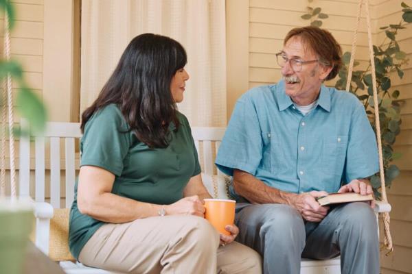A Home Instead caregiver sits on a porch swing with an elderly man, both smiling warmly, highlighting a moment of shared connection and companionship.