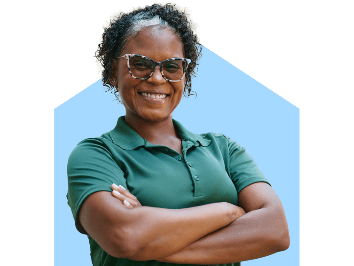 A confident Home Instead caregiver in a green uniform smiles with arms crossed, standing outdoors, representing trust and professionalism.