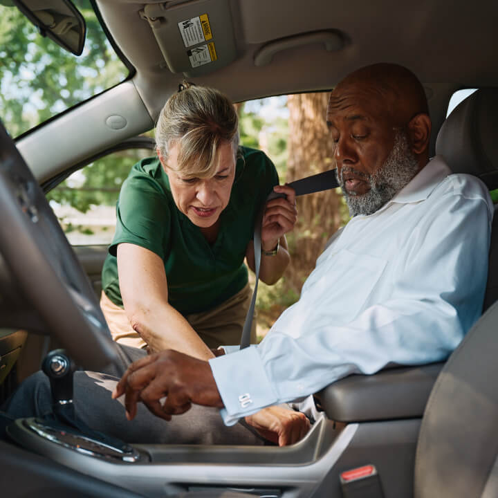 A Home Instead caregiver assists an elderly man in the passenger seat by fastening his seatbelt, ensuring safety and comfort during transportation.