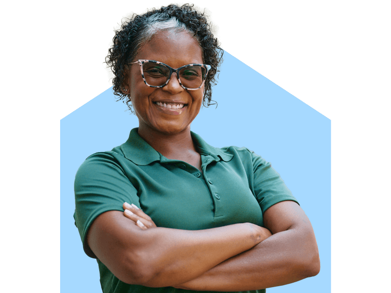 A confident Home Instead caregiver in a green uniform smiles with arms crossed, standing outdoors, representing trust and professionalism.